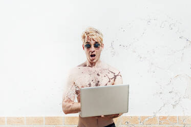 Wasser spritzt auf einen Mann ohne Hemd, der einen Laptop benutzt und an der Wand steht - JCMF01070