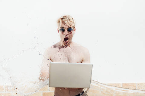 Water splashing on shirtless man wearing sunglasses using laptop against wall stock photo