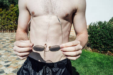 Close-up of shirtless young man holding sunglasses standing in yard during sunny day - JCMF01077