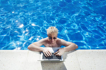 Shirtless young man wearing sunglasses using laptop on poolside during sunny day - JCMF01078