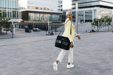 Lächelnde Geschäftsfrau mit Telefon und Tasche beim Spaziergang in der Stadt - JRFF04645