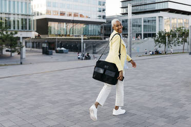 Smiling businesswoman with phone and bag walking in city - JRFF04645