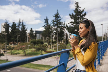 Thoughtful young woman holding drink looking away while standing by railing on footbridge - DSIF00058