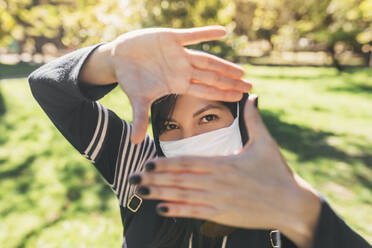 Close-up of young woman wearing mask looking through finger frames in park - DSIF00059