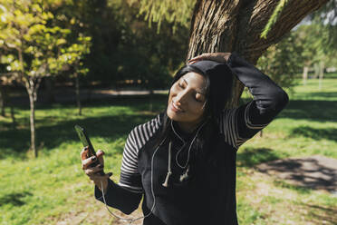 Smiling young woman taking selfie with smart phone while standing in park - DSIF00060