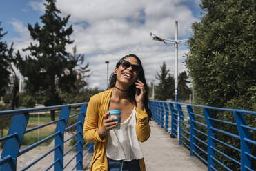 Fröhliche Frau mit Sonnenbrille, die über ihr Smartphone spricht, während sie auf einem Steg im Park steht - DSIF00063