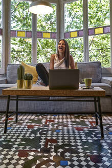 Fröhliche Frau sitzt zu Hause auf dem Sofa vor ihrem Laptop - VEGF02540
