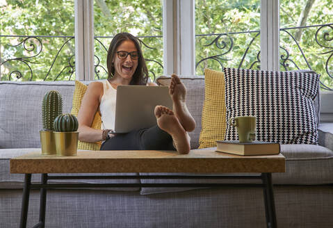 Fröhliche Frau, die einen Laptop benutzt und auf dem Sofa im Wohnzimmer sitzt - VEGF02544