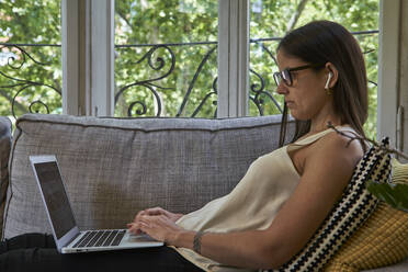 Woman wearing eyeglasses using laptop on sofa at home - VEGF02546
