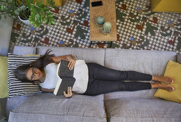 Frau liest ein Buch, während sie zu Hause auf dem Sofa liegt - VEGF02548