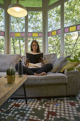 Woman reading book while sitting on sofa in living room - VEGF02552