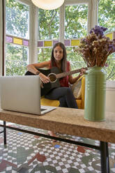 Woman learning guitar online while sitting on armchair at home - VEGF02564