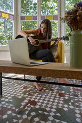 Frau lernt online Gitarre, während sie auf einem Stuhl im Wohnzimmer sitzt - VEGF02565