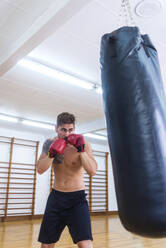 Junger bärtiger Mann trainiert in einem Fitnessstudio und schlägt mit der Faust auf seinen Boxsack ein - ADSF06722