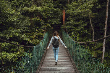 Rückenansicht junge schlanke Frau mit Rucksack geht auf Hängebrücke zu grünen exotischen Wäldern in Malaysia - ADSF06734