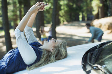 Enthusiastic woman taking self-portrait on hood of car in woods - CAIF29142