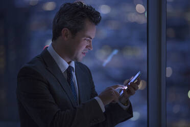 Businessman text messaging with cell phone in window at night - CAIF29227