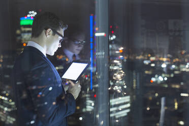 Geschäftsmann, der ein digitales Tablet in einem städtischen Fenster bei Nacht benutzt - CAIF29248