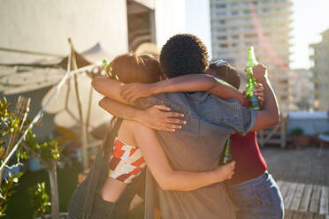 Glückliche junge Freunde umarmen sich und trinken Bier auf einem sonnigen Dach - CAIF29363