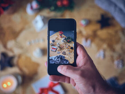 Hand eines anonymen Mannes, der ein modernes Smartphone benutzt, um verschiedene Weihnachtsdekorationen auf dem Tisch zu fotografieren - ADSF06948