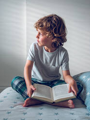 Niedlicher Junge im Schlafanzug sitzt auf einem bequemen Bett und liest ein schönes Buch. - ADSF07044