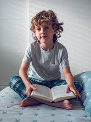 Cute boy in pajamas sitting on comfortable bed and reading nice book. - ADSF07045