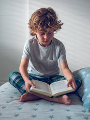 Cute boy in pajamas sitting on comfortable bed and reading nice book. - ADSF07046