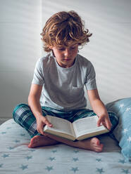 Niedlicher Junge im Schlafanzug sitzt auf einem bequemen Bett und liest ein schönes Buch. - ADSF07046