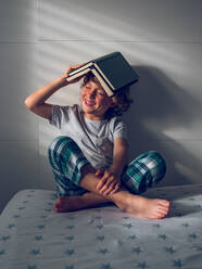 Adorable boy in pajamas wearing book on head and enjoying reading while sitting on comfortable bed. - ADSF07047