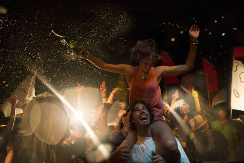 Nachtschwärmer bei einem Open-Air-Konzert, ein lächelnder Mann trägt eine Frau auf den Schultern, die Arme ausgestreckt, mit einer Bierflasche in der Hand. - MINF14649