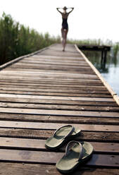 Rückansicht einer Frau im Badeanzug, die an einem Steg entlanggeht, die Hände auf dem Kopf, im Vordergrund ein Paar Flip Flops. - MINF14650