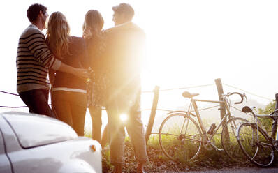 Four people standing by the side of a road, parked car and bicycles leaning against a fence, sunlight. - MINF14652