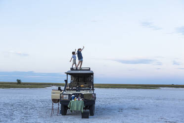 Ein Bruder und eine Schwester stehen in der Abenddämmerung in einer Salzpfannenlandschaft auf dem Dach eines Safarifahrzeugs - MINF14700