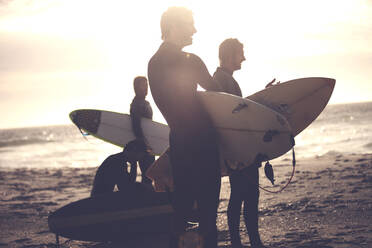 Vier Männer in Neoprenanzügen stehen an einem Sandstrand und tragen Surfbretter. - MINF14826