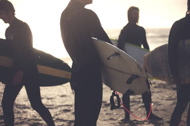 Vier Männer in Neoprenanzügen stehen an einem Sandstrand und tragen Surfbretter. - MINF14827