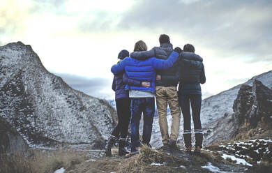 Rückansicht von vier Personen, die Arm in Arm auf einem Berg stehen, mit schneebedeckten Gipfeln in der Ferne. - MINF14831