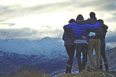 Rückansicht von vier Personen, die Arm in Arm auf einem Berg stehen, mit schneebedeckten Gipfeln in der Ferne. - MINF14833
