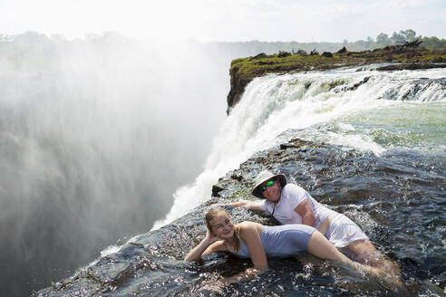 Ein Mann und ein Mädchen, Vater und Tochter, im Wasser des Devil's Pool am Rande der Victoriafälle, Nebel steigt vom fallenden Wasser auf. - MINF14879