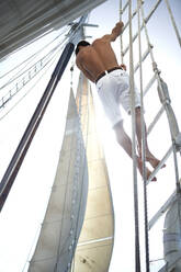 Ein Mann in weißen Shorts mit nackter Brust klettert an der Takelage eines Segelbootes hoch. - MINF14948