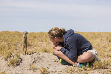 12-jähriges Mädchen, das in der Kalahari-Wüste beobachtet, wie Erdmännchen aus ihren Höhlen schlüpfen. - MINF14967