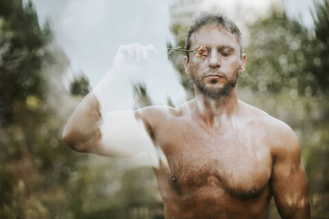 Shirtless handsome man with eyes closed holding flower on eye in forest seen through glass stock photo