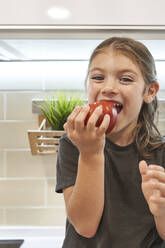 Nahaufnahme eines glücklichen Mädchens, das eine Tomate isst, während es zu Hause in der Küche sitzt - VEGF02571