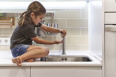 Mädchen wäscht sich die Hände im Waschbecken, während sie auf der Küchentheke zu Hause sitzt - VEGF02576