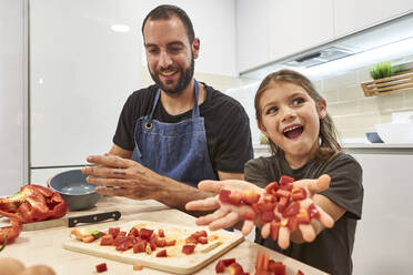 Glücklicher Vater und Tochter mit Paprikastreifen auf dem Tisch bei der Zubereitung von Speisen in der Küche - VEGF02582