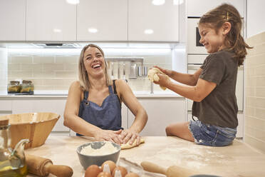 Fröhliche Mutter und Tochter kneten Pizzateig, während sie in der Küche zu Hause sitzen - VEGF02595