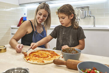 Mutter betrachtet Tochter beim Auftragen von Soße auf Pizzateig in der Küche - VEGF02611