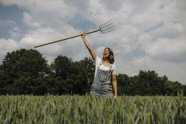 Screaming young woman lifting up hay fork in a grain field in the countryside - GUSF04203