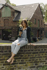 Junge Frau sitzt auf einer Backsteinmauer in einem Bauernhaus und benutzt ihr Smartphone - GUSF04225