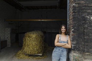 Porträt einer lächelnden jungen Frau, die sich an eine Backsteinmauer in einer Scheune auf einem Bauernhof lehnt - GUSF04234