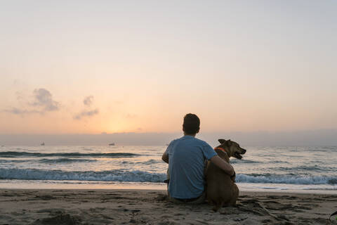 Mann sitzt mit seinem Hund und genießt die Morgendämmerung am Strand, lizenzfreies Stockfoto